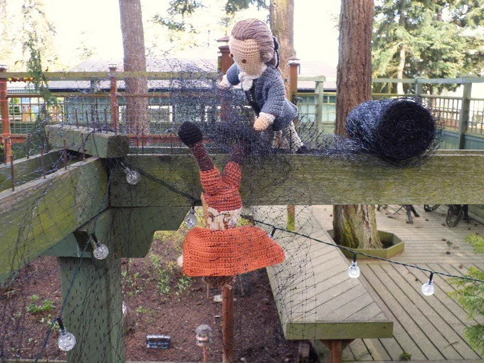 Crochet dolls of Nathaniel Sackett hanging upside down from the arbor planks, tangled in deer net while Robert Townsend tries to free him. New solar lights are hanging from the arbor & back deck in background.