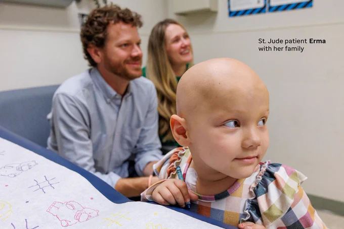 St. Jude patient Erma, photographed looking off to the side. She is photographed wearing a plaid, colorful outfit. Her family sits in the background of the image, inside a room.