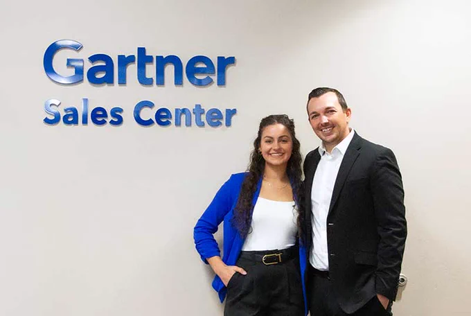 two people smile for a photo next to a logo sign that reads gartner sales center