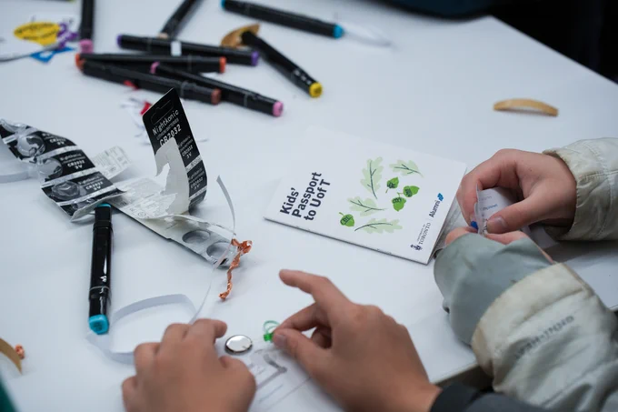 A child's hands work with a coin cell battery, copper tape and a small green LED on a white table. A folded white booklet titled "Kids' Passport to U of T" with illustrated green oak leaves and acorns sits in the middle of the table, alongside scattered black markers and a battery package.