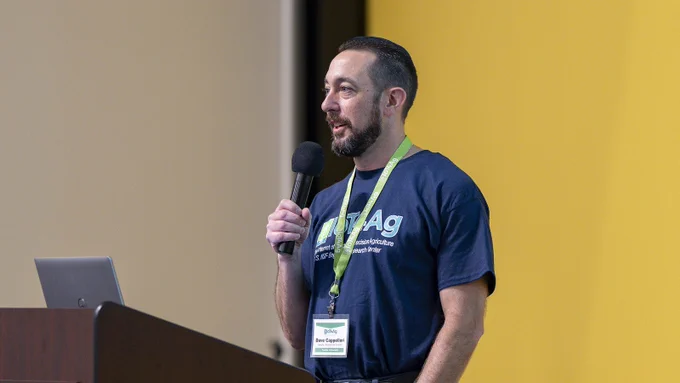 Prof. Dave Cappelleri speaks into a mic at a Purdue podium wearing an IoT4Ag t-shirt and lanyard at the 2026 Annual Retreat. 