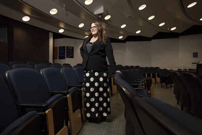 A person stands in an empty auditorium, wearing a black jacket and a polka-dotted dress. The room features rows of blue chairs and curved ceiling lights.