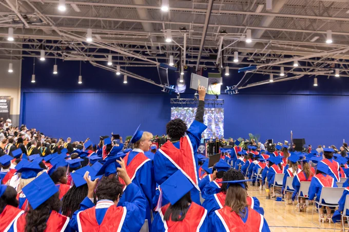 Graduation ceremony with a crowd of students dressed in blue and orange gowns and mortarboards, seated in a large indoor venue.