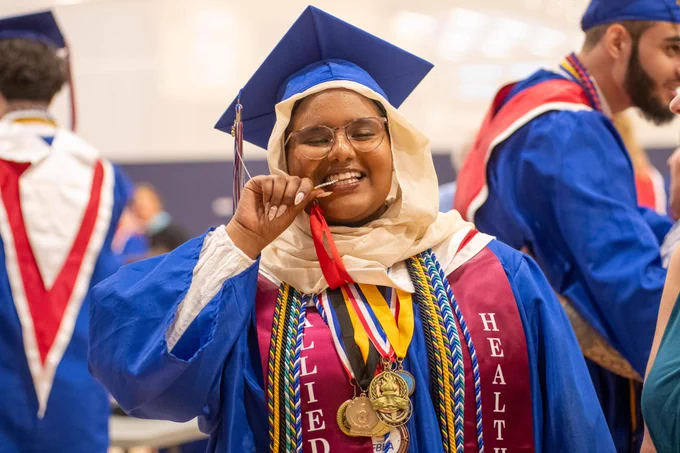 A smiling graduate in blue cap and gown holding academic medals and degrees, surrounded by fellow graduates in similar attire.