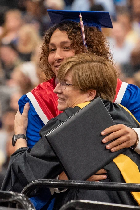 A woman in graduation regalia embracing an older woman in a wheelchair, who is also wearing graduation attire, during a graduation ceremony.