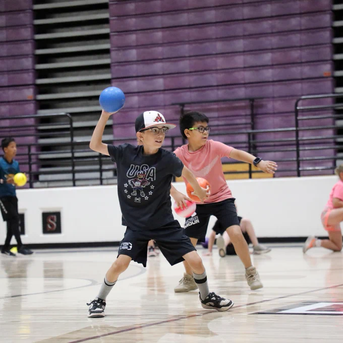 Children are playing dodgeball in a gymnasium. One child in the foreground is throwing a blue ball and wearing a hat with "USA" on their shirt. Another child nearby is holding an orange ball, ready to throw. The background shows bleachers and other participants.