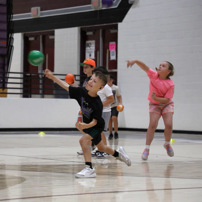 Children are actively playing dodgeball in a gymnasium. They are enthusiastically throwing and dodging colorful balls. The floor is marked with lines for sports activities, and there are doors and bleachers in the background.