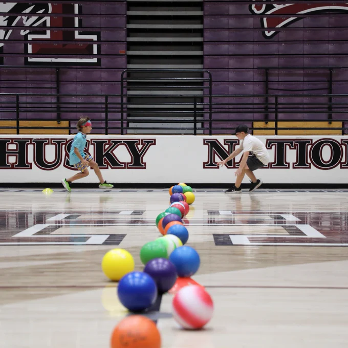A group of colorful balls is scattered on a gym floor, leading towards two children playing in the background. The wall behind them displays "Husky Nation" in large letters.