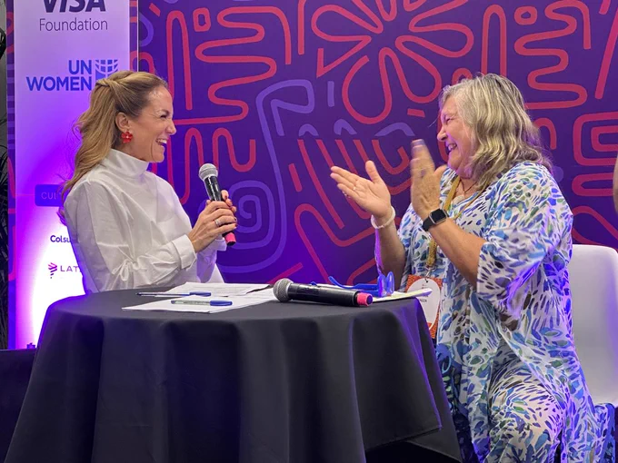 Two people are seated at a table talking into microphones. One person is smiling and holding a microphone, while the other, who is wearing a patterned outfit, is clapping. Behind them is a colorful backdrop with the logos of Visa Foundation and UN Women.