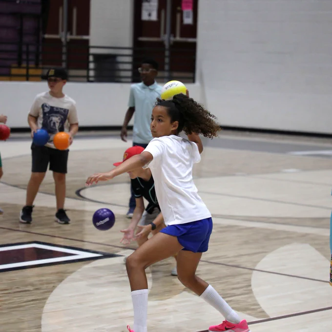 A person in a white shirt and blue shorts is playing dodgeball in a gymnasium. The gym floor is marked with lines and a visible sports logo.