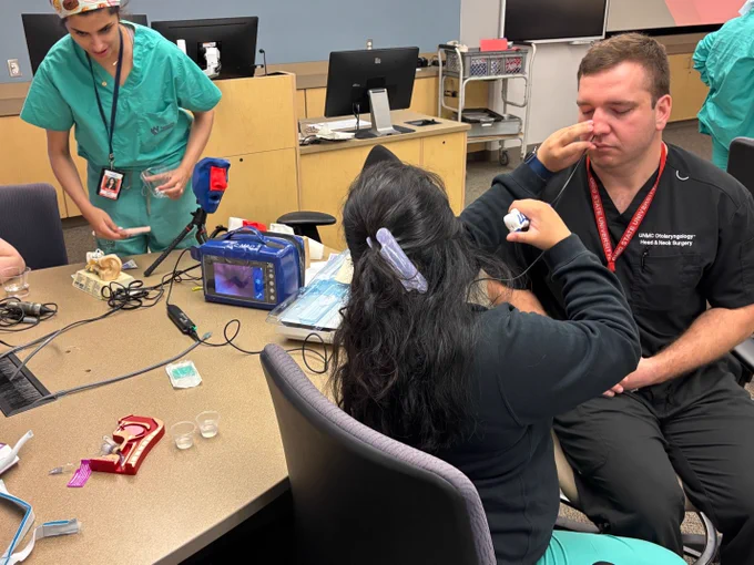ENT residents participate in a hands-on clinical skills workshop. One person performs a nasal endoscopy procedure on a seated participant while others observe and prepare equipment around a table with medical training tools and monitors.