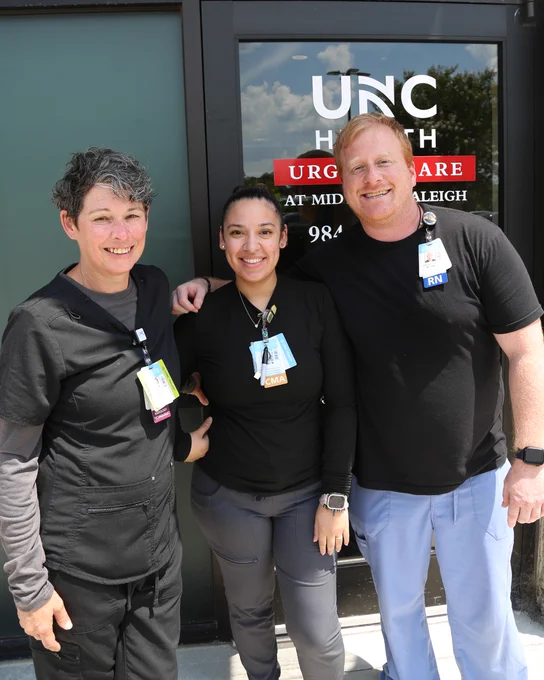 Two people are standing with arms around each other, smiling, outside a building with a UNC Health Urgent Care sign. A third person stands in front of them, also smiling.