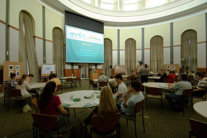 Attendees sit at round tables in a large, circular rotunda hall during the Appalachian Rural Health Institute conference. A projection screen at the front of the room displays a welcome slide introducing Dean John A. Brose, DO.
