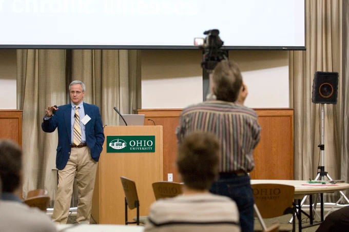 A presenter wearing a blue blazer and khaki pants stands next to an Ohio University podium, gesturing during his presentation. A video camera operator in the foreground records the presentation from the audience.