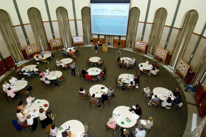 An overhead, high-angle view of the circular conference room showing numerous attendees seated at round tables. A slide titled "Activators of Stress Response" is projected on the large screen at the front of the room.