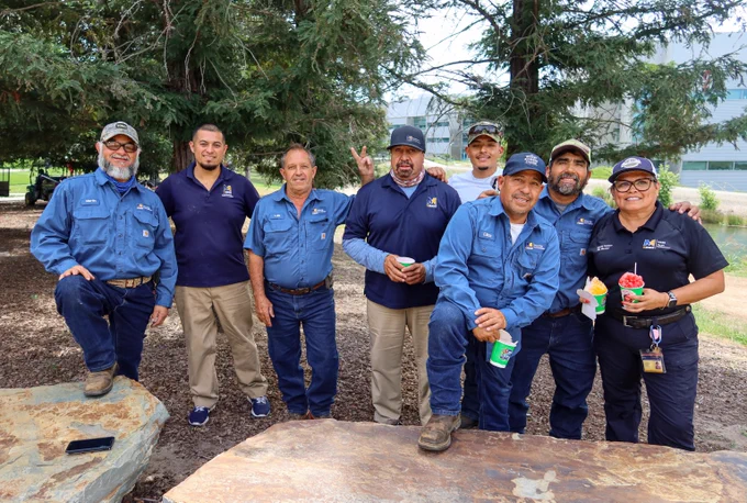 Group of eight workers in blue uniforms posing outdoors near trees, some holding colorful frozen drinks and smiling.