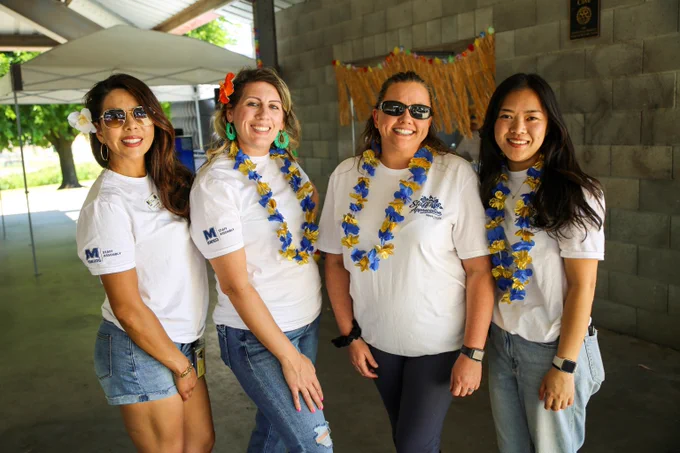 Four women wearing white event t-shirts and blue and yellow flower leis pose together under a covered outdoor area.