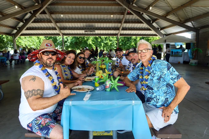 Group of people wearing leis and tropical shirts enjoying a meal together under a pavilion with festive decorations on the table.