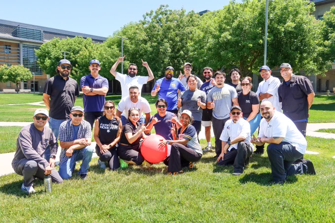 Group of diverse coworkers posing outdoors on a sunny day with a large red exercise ball on green grass.