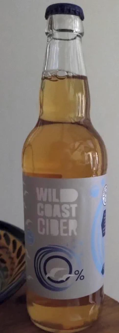 A crown-capped bottle of Wild Coast Cider 0%. The bottle's clear glass, showing a light amber coloured drink.