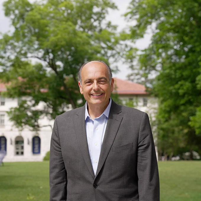 Environmental portrait of Christopher L. Augostini in a open-collar dress shirt and blazer smiling on the Emory Quad, with the Administration building and campus greenery blurred in the background