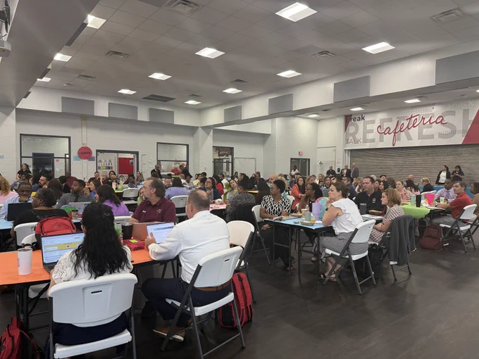 A crowded cafeteria setting with people seated at tables, some eating and conversing.
