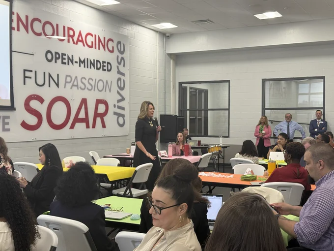 Classroom setting with people gathered around tables, large "Encouraging Fun Passion Soar" sign on wall, and some individuals engaging in discussion.