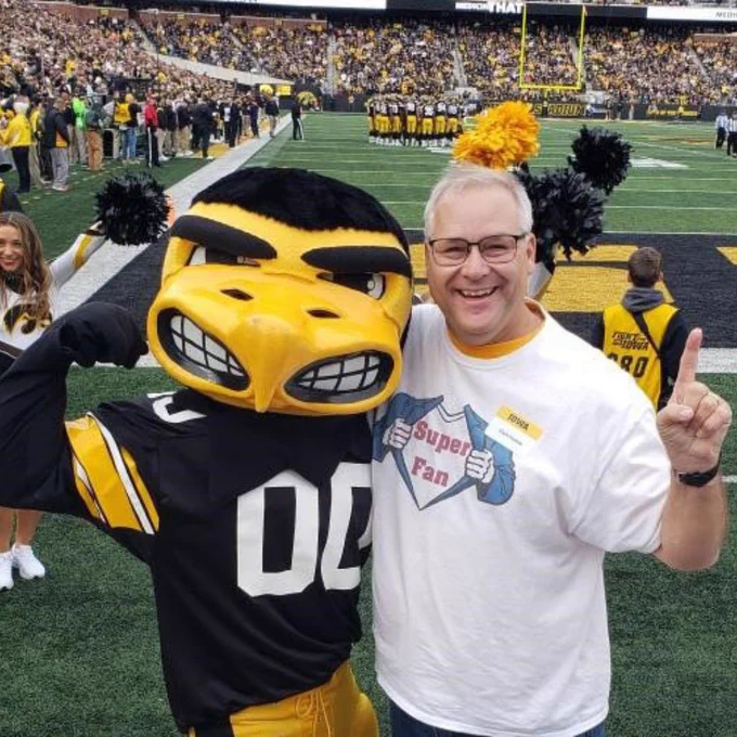 Herky the Hawk poses on the field at Kinnick Stadium with longtime Hawkeye superfan Clark Humble before an Iowa football game. Clark smiles and points upward while wearing a “Super Fan” T-shirt, with a packed stadium crowd in the background.