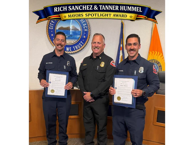 Photo of engineer Rich Sanchez and Firefighter/Paramedic Tanner Hummer standing with Huntington Beach Fire Chief Eric McCoy during a Mayor’s Spotlight Award recognition ceremony at Huntington Beach City Hall. Sanchez and Hummer hold framed certificates while a blue and gold banner above them reads “Rich Sanchez & Tanner Hummer – Mayor’s Spotlight Award.”