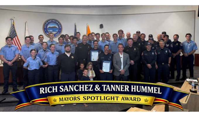 Group photo inside Huntington Beach City Hall showing Fire Explorer Advisors Rich Sanchez and Tanner Hummer standing with the Mayor, Huntington Beach Fire Department command staff, firefighters, family members, and a large group of HBFD Fire Explorers in uniform. A blue and gold banner at the bottom reads “Mayor’s Spotlight Award – Rich Sanchez & Tanner Hummer – HBFD Fire Explorer Advisors.”