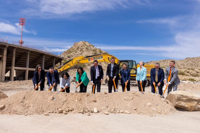 A group of people standing in a row, holding pickaxes with orange handles, are participating in a groundbreaking ceremony at a construction site on the UTEP campus. Behind them, a large "Border" branded excavator is visible. The scene is set against a backdrop of dirt, rocks, and a stadium structure with a clear blue sky above.
