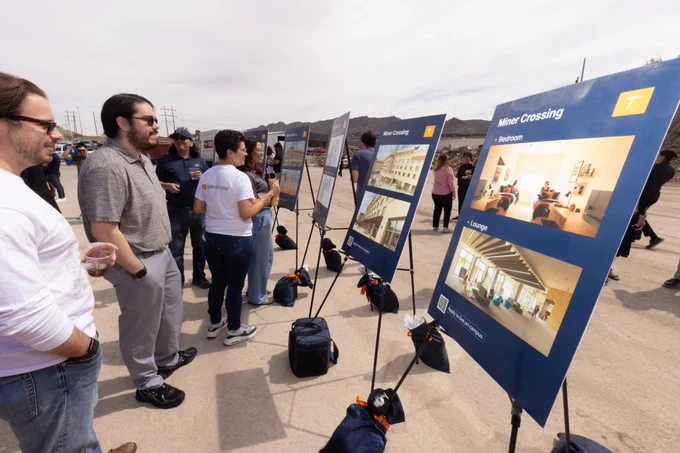 People are gathered outdoors, looking at display boards with images and information about a project called "Miner Crossing." Two display stands show interior views and labeled architectural renderings. The scene suggests a presentation or community event.