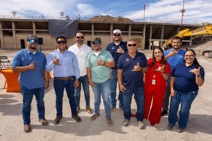A group of nine people stands outdoors in front of a stadium under construction. They are all smiling and making "picks up" signs. There is a construction vehicle and equipment in the background.