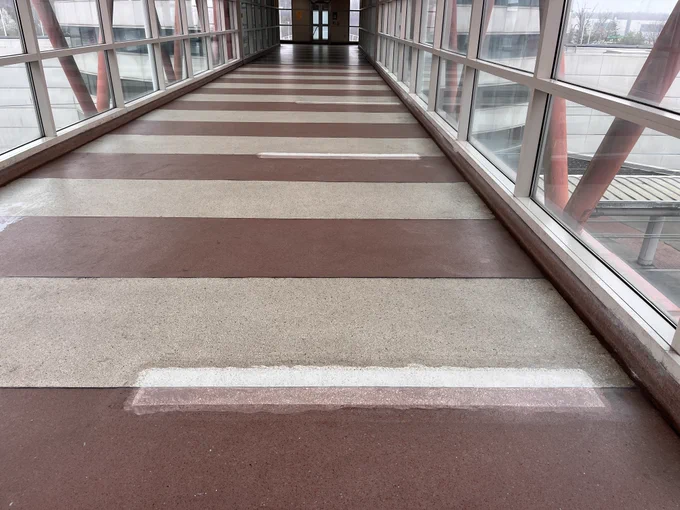 A covered pedestrian walkway with a striped floor pattern made of alternating reddish-brown and beige stripes. Large glass windows line both sides, offering a view of a modern building exterior and a glimpse of the outside. Red structural beams are visible through the glass.