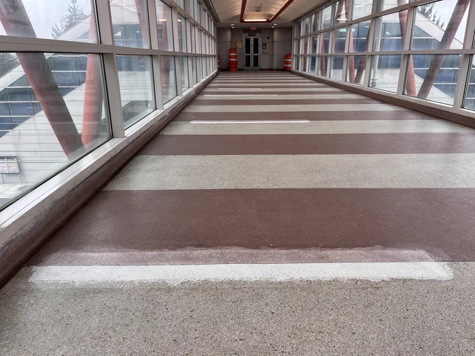 A covered pedestrian bridge with large windows on both sides, featuring a series of red and gray horizontal stripes on the floor. Two red bollards are visible near the entrance at the end of the bridge, with a closed door and EXIT sign above.