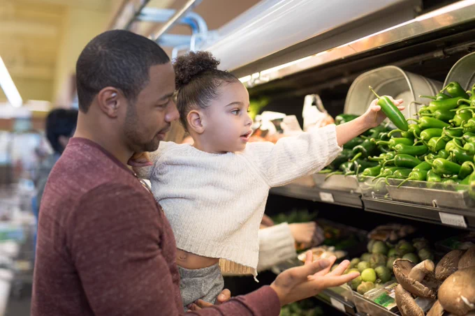 Adult and child are at a supermarket in the vegetable aisle as the child chooses a green pepper from a basket. 
