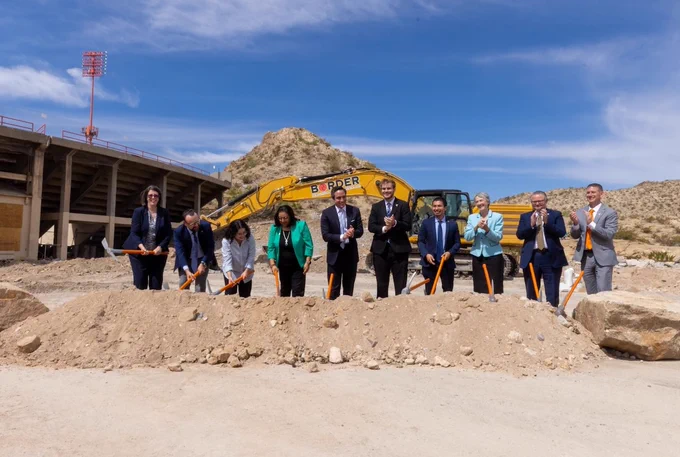 A group of people standing in a row participating in a groundbreaking ceremony at a construction site on the UTEP campus. Behind them, a large "Border" branded excavator is visible. The scene is set against a backdrop of dirt, rocks, and a stadium structure with a clear blue sky above.

