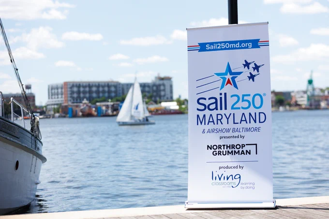 Photo of a sign that reads:

Sail250 Maryland and Airshow Baltimore, Presented by Northrop Grumman and produced by Living Classrooms.

Behind the sign is a scenic view of the Baltimore Harbor from Fells Point. 