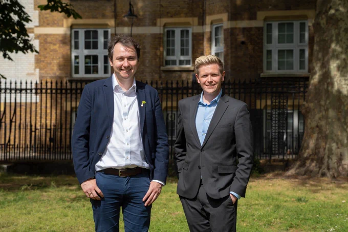 Councillors James McAsh and Victor Chamberlain stand next to each other outdoors, with railings and housing seen in the background