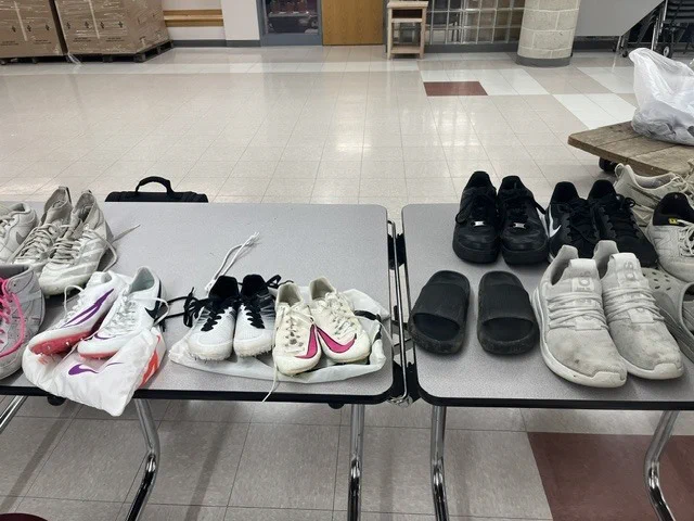 Two tables with various sneakers and footwear, including sandals, displayed in a room with white tiles and a wooden table.