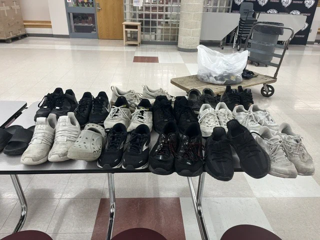 A table displays many pairs of sneakers, including black, white, and other colors, in a room with a white floor and checkered tile.