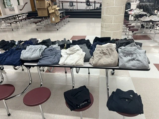 A table with folded clothing on it, with maroon stools nearby, in a room with chairs and tables.