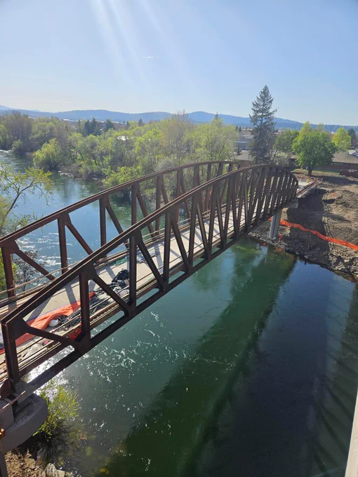 A metal pedestrian bridge spanning a river. The bridge is constructed with a series of metal trusses forming a slight arch over the water. The trusses are a rusty brown color, and the bridge has a concrete pathway. The river is calm, reflecting the bridge and surrounding greenery, with slight ripples and sparkling highlights from the sunlight. To the sides of the bridge, there is construction material visible, with some orange safety netting along the edge. In the background, dense green trees line the riverbank, and more trees and low hills extend to the horizon under a clear blue sky.
