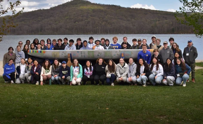 Members of Pitt's ASCE Student Chapter in front and behind the concrete canoe with a lake and hill in the background.