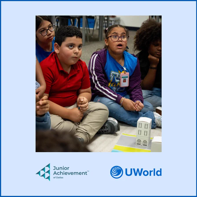 A group of young students is sitting on the floor, engaged in an activity. They are focused on a small model building in front of them. The image includes logos for Junior Achievement of Dallas and UWorld at the bottom.