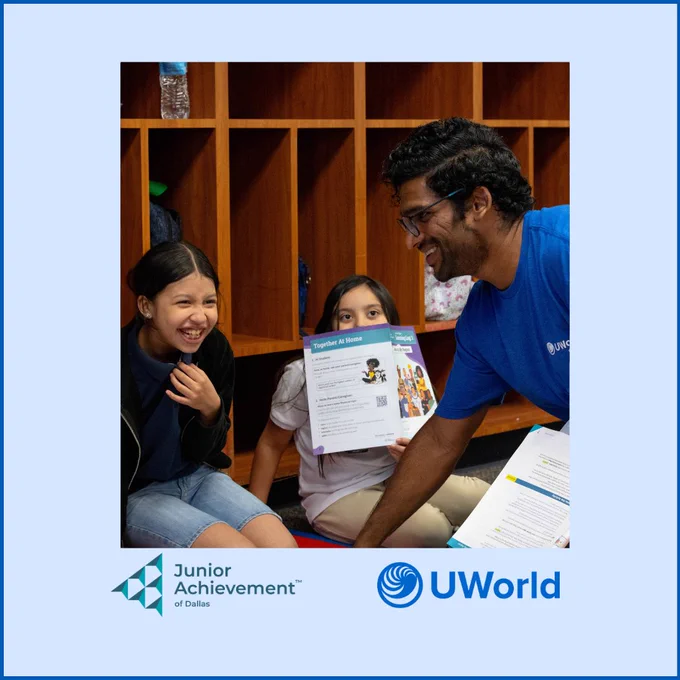 A person smiles and interacts with two children in a room with wooden cubbies. One child holds a paper with educational content. Logos for Junior Achievement of Dallas and UWorld are displayed at the bottom.