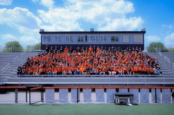Large group of people wearing orange and black seated on stadium bleachers under a blue sky with scattered clouds.