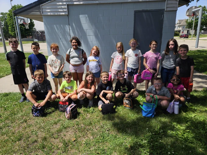 Group of children posing outdoors on grass in front of a building, some standing and others sitting with backpacks and lunch bags.