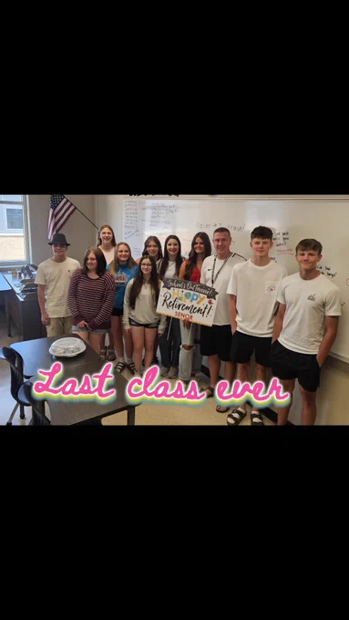 Group of students and a teacher posing in a classroom holding a retirement sign, celebrating the last class together.