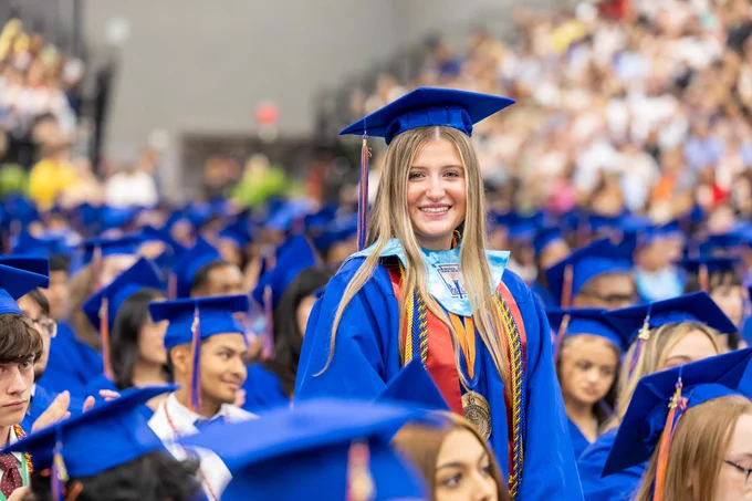 Smiling graduate in blue cap and gown standing among crowd of graduates in graduation ceremony.